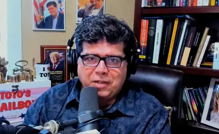 Shane Vaugh, wearing glasses and headphones, is seated in a cluttered office speaking into a microphone.