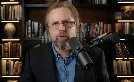 Steve Turley, a white man with red hair and beard, speaking into a microphone while seated in front of bookshelves in what appears to be a home office or library.