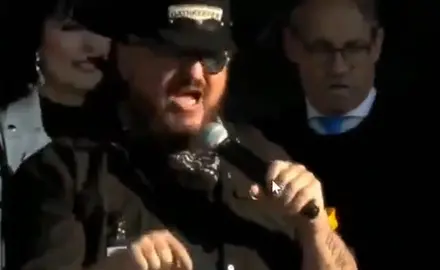 Stewart Rhodes, wearing a cowboy hat emblazoned with Oath Keepers logo," speaks into microphone held in his left hand while gesturing with his right. Rally emcee Eric Metaxas is visible behind him. 