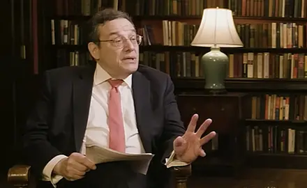 Tom Klingenstein, a white man wearing glasses and a suit and tie, holds papers in his right hand and gestures with his left. He is seated in front of bookshelves in what appears to be an office or home library. He is looking toward his left and speaking with John Eastman, who is not visible in this image cropped from video of their interview.