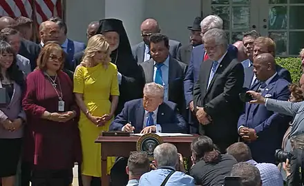 President Donald Trump seated at a desk in the White House rose garden, signing an executive order creating a Religious Liberty Commission, as White House aide Paula White and a group of religious leaders and activists look on. 