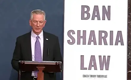 Sen. Tommy Tuberville, wearing suit and tie, stands behind lectern next to a huge poster reading BAN SHARIA LAW.