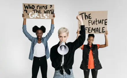 Three women, one holding a sign reading "who runs the world? Girls!", the second woman holding a megaphone, and the third holding a sign reading "the future is female."