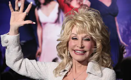 Dolly Parton, a white woman with blonde hair, wearing a white jacket and smiling and waving.
