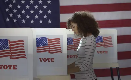 A Black woman voting at a voting booth that says "vote" on it all in front of a large American flag.