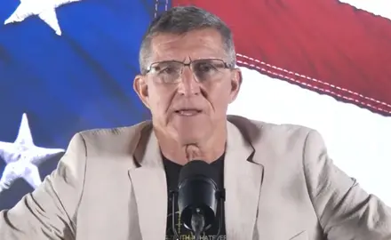 Michael Flynn speaks into a microphone seated in front of a close-up image of stars and stripes from a U.S. flag. 