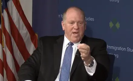Tom Homan, in suit and tie and seated in front of an American flag, gestures while speaking. 