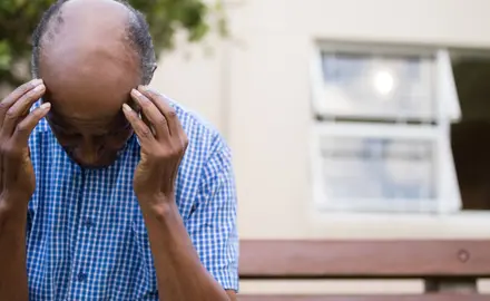 An elderly man sits on a bench and holds his head in his hands.