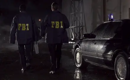 Two men in FBI jackets walk past a black car at night.