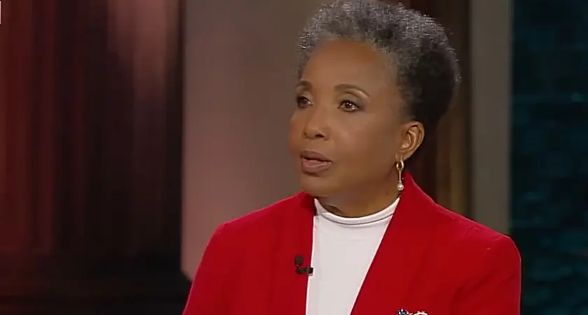 Carol Swain, a Black woman with graying hair and wearing a white shirt under a bright red jacket, is seated on set and looking toward her off-camera interviewer