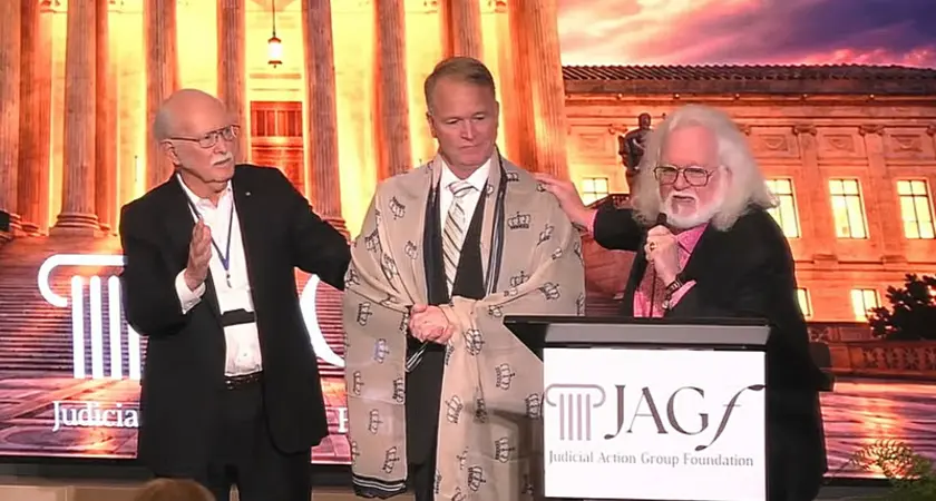 Dominionist Chuck Pierce, who has a gray beard and long gray hair, stands behind a lectern with the logo for the Judicial Action Group Foundation logo. His right hand rests on the shoulder of Phillip Jauregui, who has a shawl embroidered with crowns draped over his shoulders. A third man has his hand up in prayer. They are standing before an image of the U.S. Supreme Court building lit up at night. 