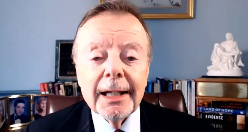 Gary Bauer, in suit and tie, is seated in what appears to be a home office. Visible behind him are some books and photos and a small model of what appears to be Michelangenlo's Pietà