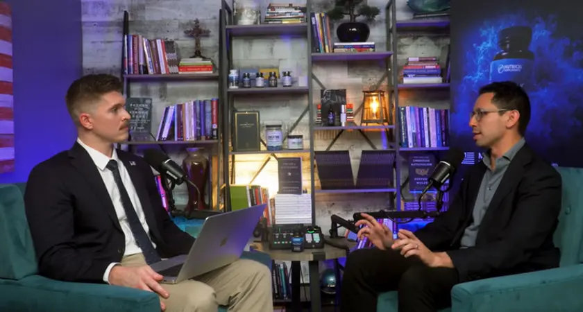 Joshua Haymes and James Fishback are seated in arm chairs facing each other in front of a set of bookshelves. Haymes has a laptop open on his lap.