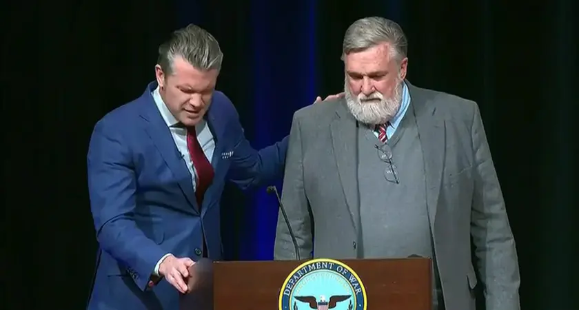 Defense Secretary Pete Hegseth and Christian nationalist pastor Doug Wilson praying at lectern bearing the logo of the "Department of War." Their heads are bowed, and Hegseth has one hand on the lectern and one on Wilson's shoulder.
