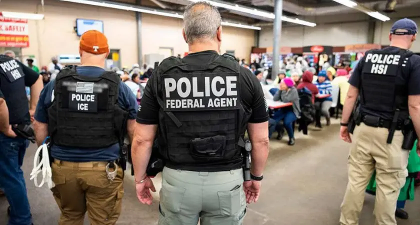 Photo shows several officers identified as police and/or ICE from the back as they face workers gathered in what appears to be a workplace lunchroom
