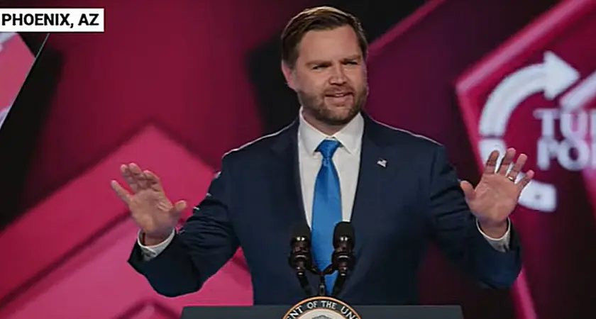 Vice President J.D. Vance gestures with his hands while speaking at lectern on stage at TPUSA's AmericaFest; the TPUSA logo is partially visible behind him.