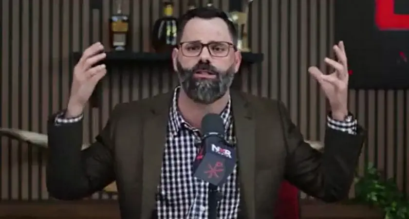 Joel Webbon gestures with both hands while seated behind a microphone that displays the logo for his NXR (New Christian Right) Studios