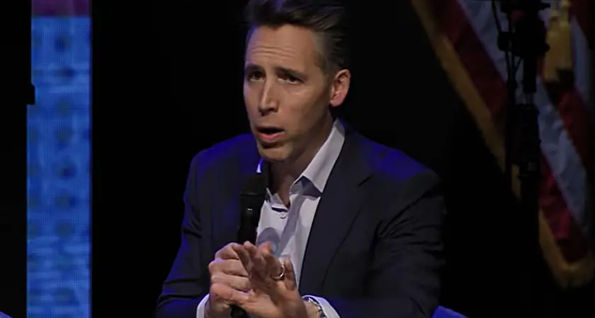 Sen Josh Hawley holds a microphone in his right had and gestures toward the audience with his left hand while seated on stage