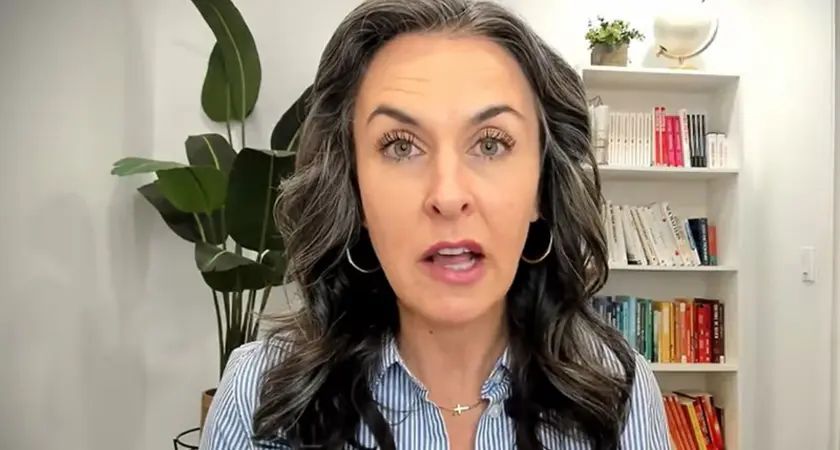 Katy Faust, a white woman with long black hair streaked with gray, speaks into the camera; a bookshelf and plant are visible behind her