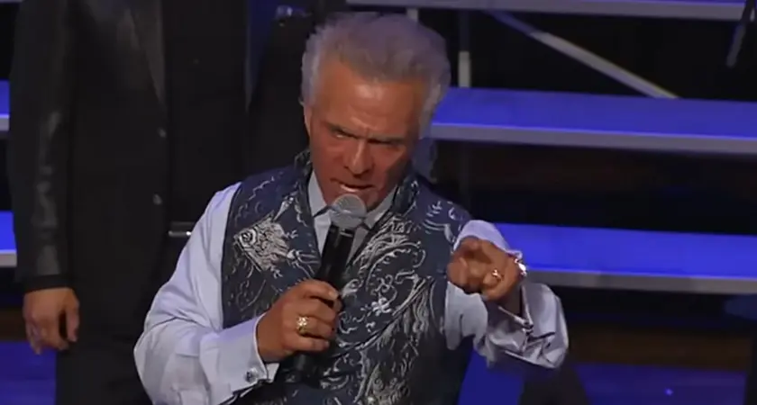 Kent Christmas, standing on stage at the Ryman Auditorium, speaks into a microphone in his right hand while pointing toward the crowd with his left hand; he looks angry.