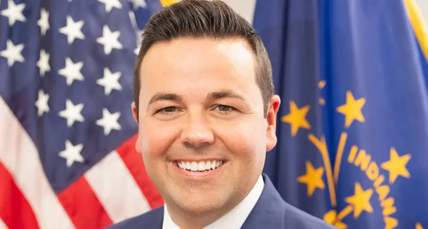 Portrait-style official photo of Indiana Lt. Gov. Micah Beckwith smiling into the camera with U.S. and Indiana flags visible behind him.