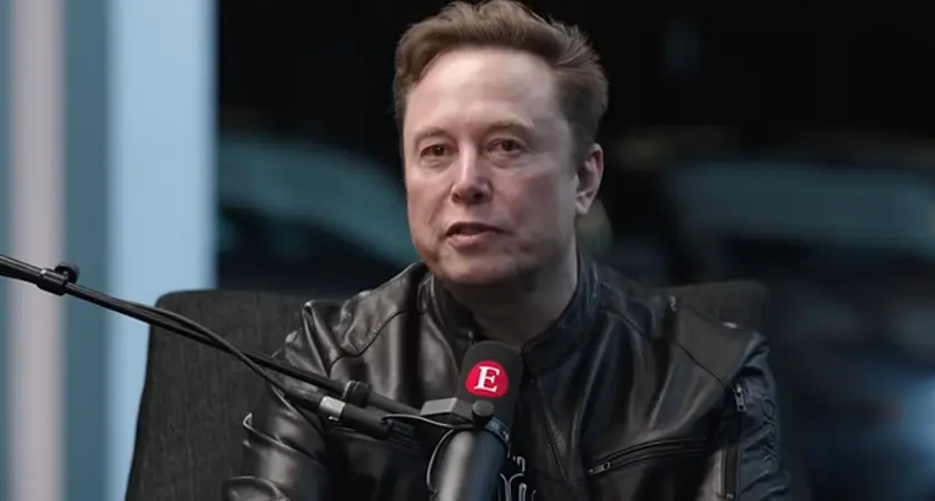 Elon Musk, in black shirt and leather jacket, is seated at a microphone that is emblazoned with a capital letter E, the logo for The Economist magazine
