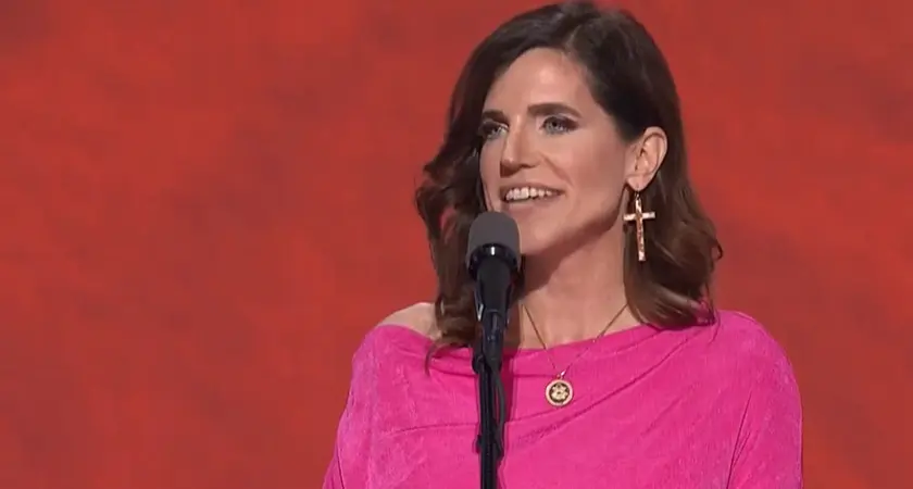 Rep. Nancy Mace, wearing bright pink dress and oversize cross-shaped earrings, speaks into a microphone in front of a red background on stage at the 2024 Republican National Convention.