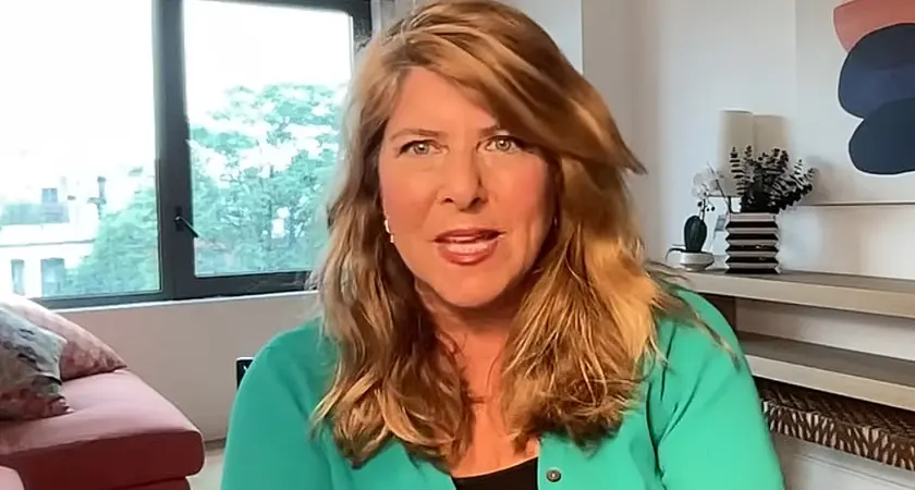 Naomi Wolf is seated in what appears to be a home office, with a window, shelf, and art visible behind her.