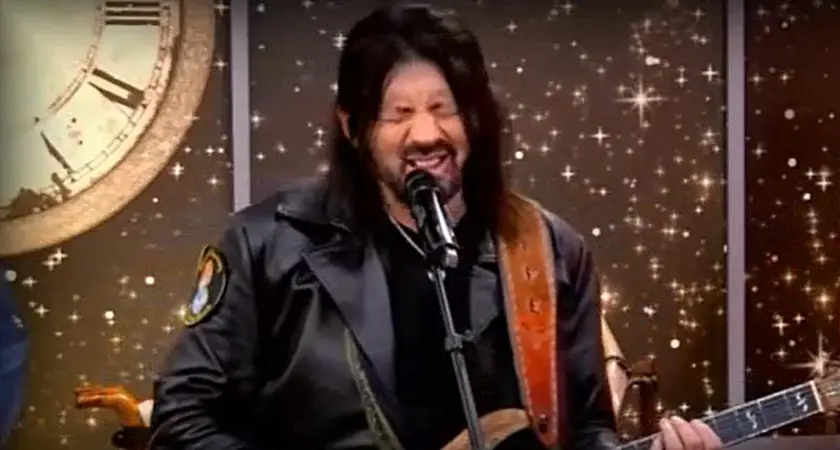 Robin Bullock, a white man with long black hair, grimace while speaking/singing into a microphone and holding a guitar. 