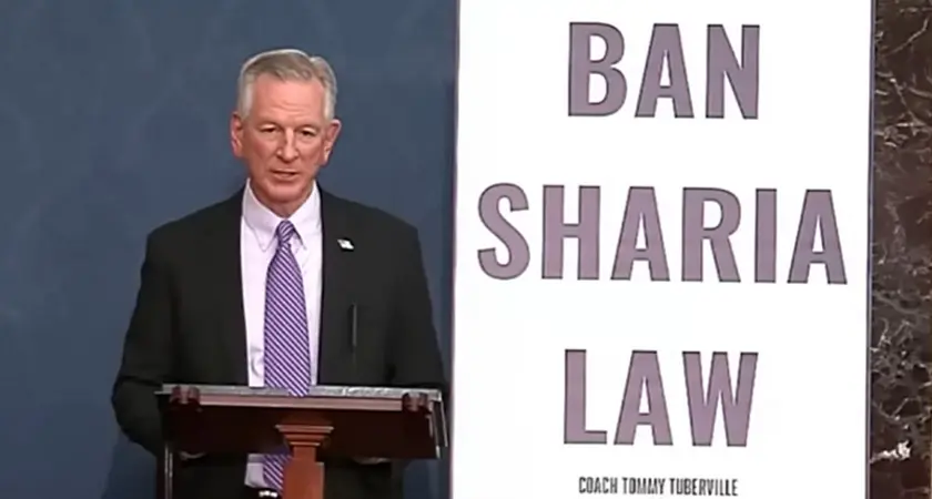 Sen. Tommy Tuberville, wearing suit and tie, stands behind lectern next to a huge poster reading BAN SHARIA LAW.