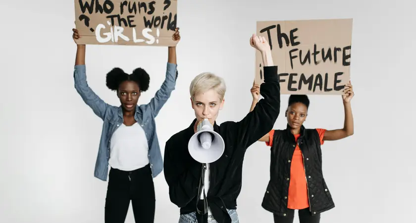Three women, one holding a sign reading "who runs the world? Girls!", the second woman holding a megaphone, and the third holding a sign reading "the future is female."