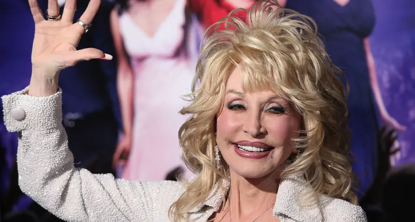 Dolly Parton, a white woman with blonde hair, wearing a white jacket and smiling and waving.