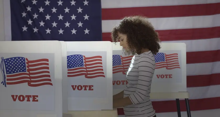 A Black woman voting at a voting booth that says "vote" on it all in front of a large American flag.
