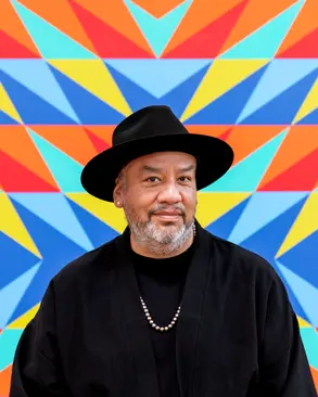 A photo of Jeffrey Gibson, a Black man wearing a black hat and black shirt standing in front of a multi-colored wall. 