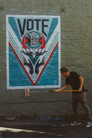 Artist Shepard Fairey wheatpasting a poster to a wall. the poste reads "Vote"