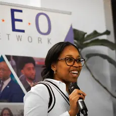 Woman speaking into a microphone in front of a sign that says YEO Network