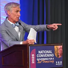 Bill Nye wearing a gray suit and blue bow tie standing at a podium at the Young Elected Officials Network Convening 2026 and holding a copy of the Constitution. 