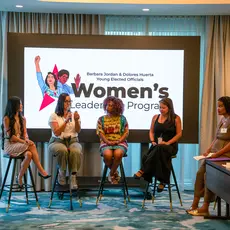 Five women sit on stools in front of a sign that reads "Women's Leadership Program"