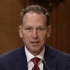 Brian Burch, a white man with receding hairline wearing blue suit and red tie seated at a table at his confirmation hearing.