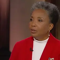 Carol Swain, a Black woman with graying hair and wearing a white shirt under a bright red jacket, is seated on set and looking toward her off-camera interviewer