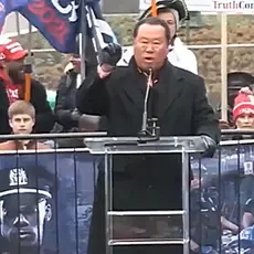 Che Ahn speaking at microphone at outdoors Jan. 5, 2021 rally on Freedom Plaza near the White House. Ahn is wearing black coat and gloves, people holding Trump flags are visible behind him.