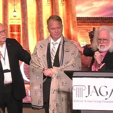 Dominionist Chuck Pierce, who has a gray beard and long gray hair, stands behind a lectern with the logo for the Judicial Action Group Foundation logo. His right hand rests on the shoulder of Phillip Jauregui, who has a shawl embroidered with crowns draped over his shoulders. A third man has his hand up in prayer. They are standing before an image of the U.S. Supreme Court building lit up at night. 