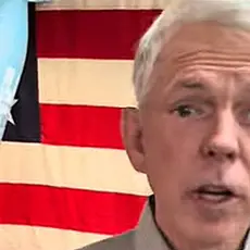 David Barton speaks into the camera; visible behind him is a World War II era (?) bomb hanging in front of a U.S. flag.