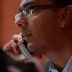 Close up side-angle view of Dinesh D'Souza looking serious and looking off camera to the left while holding a cell phone to his right ear.