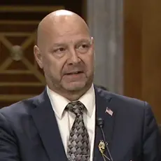Doug Mastriano in suit and tie is seated at a table in a Senate hearing room, speaking to an off-camera senator