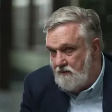 Douglas Wilson, a white man with gray hair and beard, is looking off camera to the CNN reporter interviewing him. The background is blurred. 