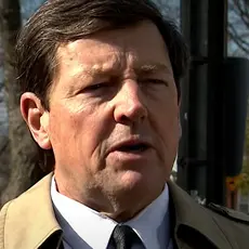 Ed Martin, wearing suit and tie and tan overcoat, is being interviewed outdoors; visible behind him are neighborhood homes and trees. 