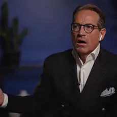 Metaxas gestures with both arms open while sitting in what appears to be a living room or home office