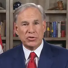 Gov. Greg Abbott wearing blue suit and red tie speaks to the camera; bookshelves are visible behind him with an American flag partially visible to the left and Texas flag to the right