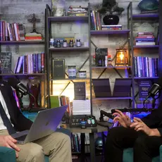 Joshua Haymes and James Fishback are seated in arm chairs facing each other in front of a set of bookshelves. Haymes has a laptop open on his lap.